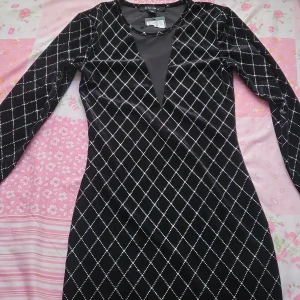 Svart klänning med rutmönster - Snygg svart klänning med ett elegant rutmönster med strass. Klänningen har en v-ringad design med meshdetalj och långa ärmar. Perfekt för en kväll ute eller en speciell tillställning.