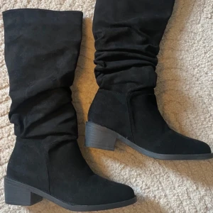 Svarta stövlar med klack - Snygga svarta stövlar i mocka med en bekväm klack. De har en stilren design med rynkade detaljer längs skaftet. Perfekta för både vardag och speciella tillfällen.