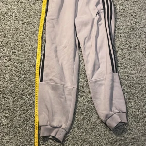 Grå mjukisbyxor från Adidas - Säljer ett par grå mjukisbyxor från Adidas med klassiska svarta ränder längs sidorna. Byxorna har en elastisk midja och muddar vid bensluten för en bekväm passform. Perfekta för en avslappnad stil.