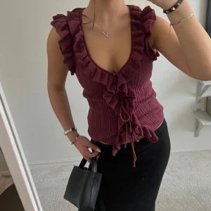 Vinröd Zara topp - Snygg vinröd ribbad topp med volangdetaljer och knytning framtill. Toppen har en v-ringad design och är ärmlös, vilket gör den perfekt för varmare dagar.