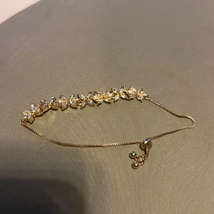 Guldarmband med blommotiv - Elegant guldarmband med blommotiv och gnistrande stenar. Armbandet har en justerbar kedja med små kulor i slutet för en perfekt passform. Perfekt för att lägga till en touch av glamour till din outfit.