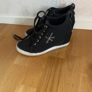 Svarta sneakers med kilklack från Calvin Klein - Snygga svarta sneakers med kilklack från Calvin Klein. Skorna har snörning och en vit sula som ger en sportig look. Perfekta för dig som vill ha både stil och komfort.