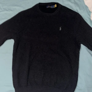Mörkgrå stickad tröja från Polo Ralph Lauren - Säljer en stilren mörkgrå stickad tröja från Polo Ralph Lauren med det klassiska broderade logotypen på bröstet. Tröjan har en rund halsringning och långa ärmar, perfekt för kyligare dagar. Passar både till jeans och chinos för en avslappnad look.