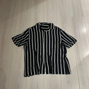 Svart och vit randig skjorta - Snygg svart skjorta med vita ränder och korta ärmar. Perfekt för en avslappnad stil. Skjortan har knappar framtill och en klassisk krage ✅