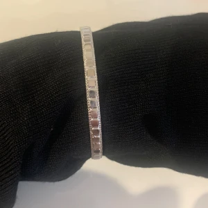 Silverfärgat armband - Elegant och stilrent armband i av metall i silverfärg med en fyrkantig mönsterdesign. Armbandet är öppet i ändarna vilket gör det lätt att justera för en bekväm passform. Perfekt för att lägga till en subtil glans till din outfit.
