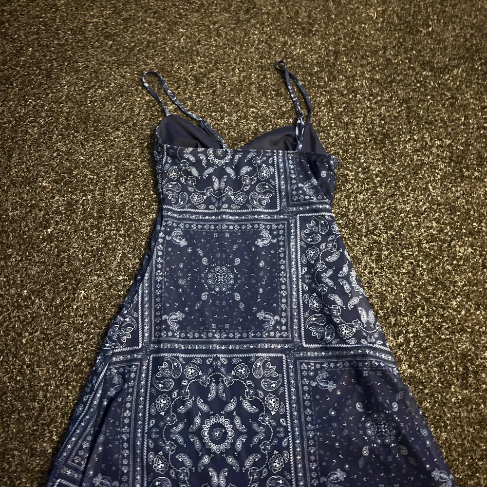 Snygg blå klänning med ett detaljerat mönster och smala axelband. Klänningen har en knytning framtill och en lätt utställd form. Perfekt för en avslappnad sommarlook.. Mekot.