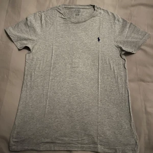 Grå t-shirt från Polo Ralph Lauren - Säljer en klassisk grå t-shirt från Polo Ralph Lauren med korta ärmar. Den har en liten broderad logga i mörkblått på bröstet. Perfekt för en avslappnad stil.