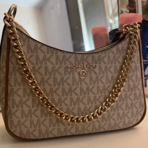 Beige axelväska från Michael Kors - Snygg beige axelväska från Michael Kors med det klassiska MK-mönstret. Väskan har en guldfärgad kedja och dragkedja upptill. Perfekt för att ge din outfit en lyxig touch.