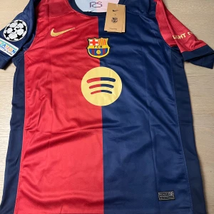 FC Barcelona fotbollströja - Snygg FC Barcelona tröja i rött och blått med klubbmärke och Nike-logga. Kortärmad design med Champions League-märke på ärmen och spelarnamn och nummer 19 på ryggen. Perfekt för fans!
