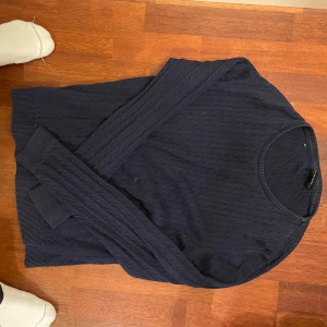 Mörkblå stickad tröja - Säljer en stilren mörkblå stickad tröja med ett diskret kabelmönster. Tröjan har långa ärmar och ribbade muddar vid ärmslut och nederkant. Perfekt för kyligare dagar.