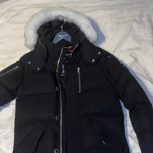 Svart dunjacka från Moose Knuckles - Säljer en svart dunjacka från Moose Knuckles med en snygg pälsfodrad huva. Jackan har dragkedja och knappar framtill samt flera praktiska fickor. Perfekt för kalla vinterdagar.