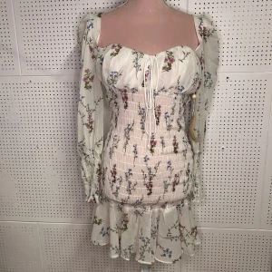 Blommig klänning med volangdetaljer - Söt blommig klänning från For Love & Lemons i vitt med rosa och blå blommor. Klänningen har långa ärmar och en smickrande smockad midja. Den har en knytning framtill och volangdetaljer vid fållen. Perfekt för en romantisk look!