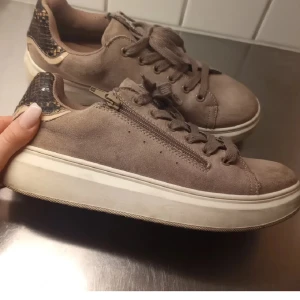 Bruna sneakers med ormskinnsdetaljer - Snygga bruna sneakers med ormskinnsdetaljer på hälen. Skorna har snörning och en dragkedja på sidan för enkel på- och avtagning. Perfekta för en stilren look. De är ifrån VOX (vegan shoes). 