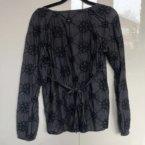 Grå blus med spetsdetaljer - Snygg grå blus med blommigt mönster och spetsdetaljer. Blusen har långa ärmar och ett knytband i midjan för en fin passform. Perfekt för en stilren look.