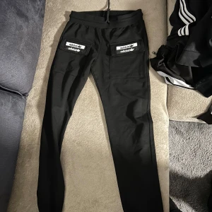 Svarta mjukisbyxor från Adidas - Snygga svarta mjukisbyxor från Adidas med vita logotyper på fickorna. Byxorna har en bekväm passform och är perfekta för en avslappnad stil. De har elastisk midja och är tillverkade i ett mjukt material.