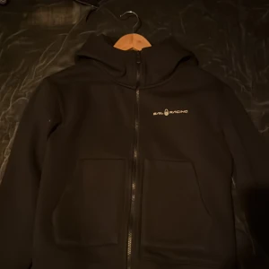 Svart jacka från Sail Racing - Säljer en svart jacka från Sail Racing med dragkedja och huva. Jackan har en stilren design med logotyp på bröstet och baksidan av huvan. Perfekt för en casual look.