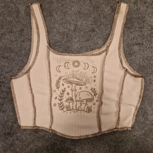 Beige ribbad linnetopp med tryck - Säljer en beige ribbad linnetopp med ett  tryck av svampar och månfaser på framsidan. Toppen har breda axelband och en croppad passform. Perfekt för en avslappnad stil.