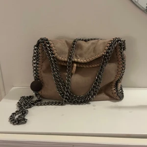 Stella McCartney liknande väska - Snygg rosa axelväska med coola kedjedetaljer längs kanterna och som axelrem. Väskan har en unik design med synliga sömmar och en rund metallbricka som detalj. Perfekt för att ge din outfit en edgy touch. En av ”sömmarna” har åkt upp men inget man tänker på så mycket!