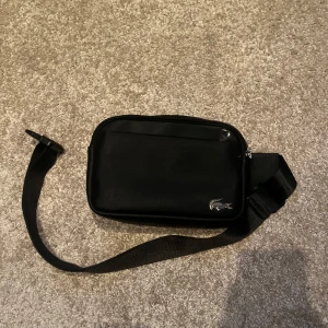 Svart axelväska från Lacoste - Snygg svart axelväska från Lacoste med justerbar rem och dragkedja. Perfekt för att bära med sig det viktigaste. Den har en stilren design med Lacoste-loggan i metall på framsidan.