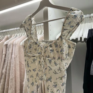 Blommig topp med knytning från Bershka  - Söt blommig topp från Bershka med knytning framtill. Toppen har breda axelband och ett romantiskt blommönster i blått och grönt på en vit bakgrund. Perfekt för en somrig look!