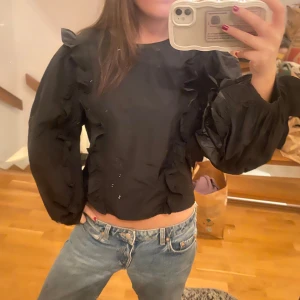 Svart blus med volangdetaljer - Säljer en svart blus med volangdetaljer på ärmarna. Blusen har långa ärmar och en croppad passform. Perfekt för att ge en stilfull touch till din outfit. passar xs- m