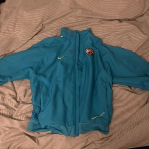 Turkos jacka från Nike med FCB-logga - Säljer en turkos jacka från Nike med FC Barcelona-logga på bröstet. Jackan har dragkedja och långa ärmar, perfekt för sportiga tillfällen. Den har även gula detaljer och är tillverkad i ett lätt syntetmaterial.