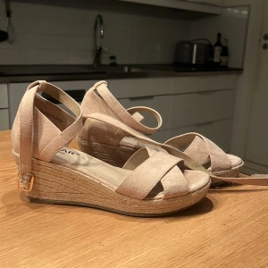 Beige kilklackssandaler från Linear - Snygga beige sandaler med kilklack och korsade remmar från Linea. Sandalerna har en justerbar rem runt ankeln med ett guldfärgat spänne. Perfekta för sommarens alla tillfällen.