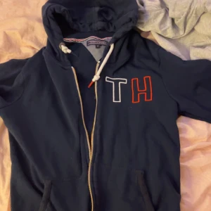 Mörkblå hoodie från Tommy Hilfiger - Säljer en snygg mörkblå hoodie från Tommy Hilfiger med dragkedja och huva. Den har broderade bokstäver 'TH' i vitt och rött på bröstet. Perfekt för en avslappnad stil.
