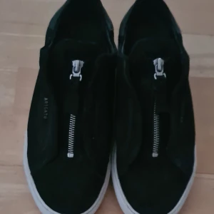 Svarta sneakers från Axel Arigato - Snygga svarta sneakers från Axel Arigato med dragkedja på ovansidan. Skorna har en stilren design med vit sula och är tillverkade i mocka. Perfekta för en trendig look.