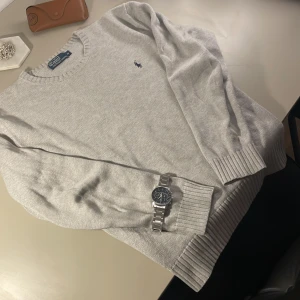 Grå stickad tröja från Ralph Lauren - Säljer en stilren grå stickad tröja från Ralph Lauren med det klassiska logotypbroderiet på bröstet. Tröjan har långa ärmar och ribbade muddar vid ärmslut och nederkant. Perfekt för en avslappnad men ändå elegant look.