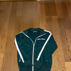 Mörkgrön jacka från Palm Angels - Palm Angels Track Jacket 	•	Storlek: S 	•	Färg: Grön med vita detaljer 	•	Nypris: Cirka 4000 kr 	•	Autentisering: Äkthetsintyg finns via QR-koden (Certilogo) 	•	Skick: Gott skick, använd få gånger  Snygg och tidlös track jacket från Palm Angels, perfekt för både vardag och mer uppklädda tillfällen. Vid fler frågor eller bud, skriv gärna! 😀