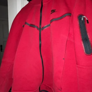 Röd fleecejacka från Nike - Säljer en röd fleecejacka från Nike med svarta dragkedjor och detaljer. Jackan har en normal passform och är perfekt för kyligare dagar. Den har långa ärmar och en praktisk ficka på ärmen.