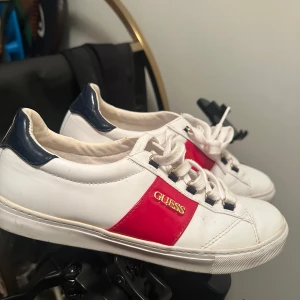 Vita sneakers från Guess med röd detalj - Snygga vita sneakers från Guess med en röd detalj och märkeslogga på sidan. Skorna har snörning och en kontrasterande mörkblå häl. Perfekta för en stilren look. Pris kan diskuteras vid snabbaffär.