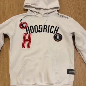 Vit dress från Hoodrich - Säljer en vit dress från Hoodrich med coola detaljer som broderade märken och text på framsidan. Hoodien har en klassisk känguruficka och en bekväm huva. Perfekt för en avslappnad stil.(hela dressen)