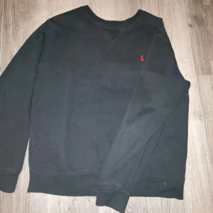 Svart tröja från Ralph Lauren - Säljer en klassisk svart tröja från Ralph Lauren med det ikoniska röda logotypbroderiet på bröstet. Tröjan har långa ärmar och en rund halsringning. Perfekt för en stilren look.