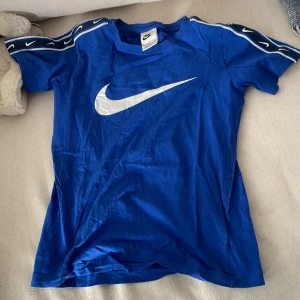 Blå t-shirt från Nike - Säljer en blå t-shirt från Nike med det ikoniska swoosh-loggan i vitt på bröstet. T-shirten har korta ärmar med vita detaljer längs axlarna. Perfekt för en sportig look eller träning.