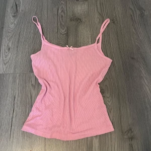 Rosa ribbad linnetopp från H&M Divided - Säljer en söt rosa ribbad linnetopp från H&M Divided. Toppen har smala axelband och en liten rosett framtill. Perfekt för varma dagar eller som ett lager under en tröja.