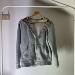 Grå hoodie från burberry - Säljer en stilren grå hoodie med dragkedja och ett klassiskt rutigt foder i huvan. Tröjan har långa ärmar och praktiska fickor framtill. Perfekt för en avslappnad stil.