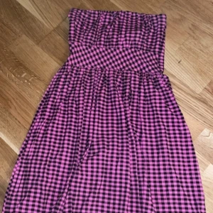 Rutig axelbandslös klänning - Säljer en söt axelbandslös klänning med rosa och svart rutigt mönster. Perfekt för sommarens varma dagar. Klänningen har en smickrande passform och är lätt att bära.