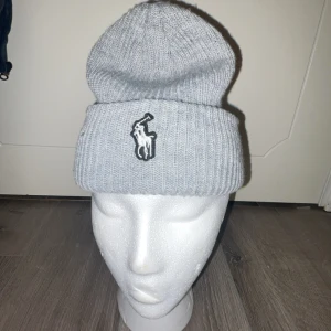Grå stickad Ralph laruen mössa - Snygg grå stickad mössa med en broderad logga framtill. Perfekt för att hålla värmen under kyliga dagar. Den ribbade designen ger en klassisk look.