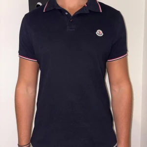 Svart pikétröja från Moncler - Snygg svart pikétröja från Moncler med klassisk krage och knappar. Tröjan har röda och vita detaljer på krage och ärmslut samt Moncler-logga på bröstet. Perfekt för en stilren look. Storlek L men sitter som M, då jag alltid bär M. Som ni kan se på bilden.  Knappt använd då jag fick en annan i present, den är som ny. Köpt för 3 500 på NK