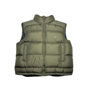 Vintage grön Nike puff jacka 167cm lång - Säljer en grön dunväst från Nike med dragkedja och hög krage. Västen har en diskret logga på bröstet och är perfekt för kyligare dagar. Den är quiltad för extra värme och stil.