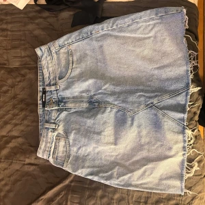 Blå jeanskjol med fransar - Säljer en cool blå jeanskjol med fransig nederkant. Kjolen har en klassisk femficksdesign och en rå kant som ger en avslappnad look. Perfekt för en trendig stil!