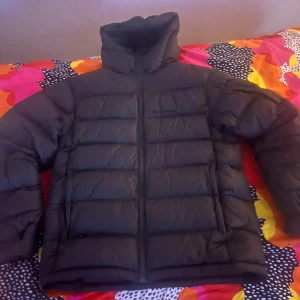 Svart pufferjacka från Peak Performance - Säljer en svart pufferjacka från Peak Performance. Jackan har en quiltad design med dragkedja framtill och en hög krage för extra värme. Perfekt för kyliga dagar och håller dig varm och bekväm. En stilren och funktionell jacka för vintern.