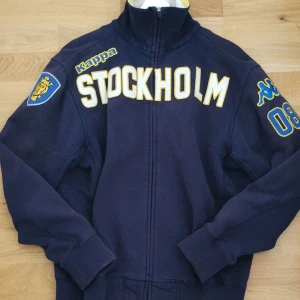Kappa tracktop Stockholm - Snygg blå jacka från Kappa med stort Stockholm-tryck i gult på framsidan. Jackan har långa ärmar och dragkedja framtill. Den är dekorerad med broderade detaljer och märken på ärmarna. Färgen är blekt på sina ställen.