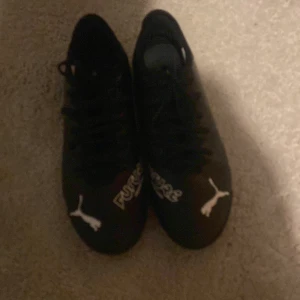 Svarta fotbollsskor från Puma - Säljer ett par svarta fotbollsskor från Puma med snörning och mönstrad sula för bra grepp. Skorna har en stilren design med Puma-loggan i vitt på sidan och framtill.