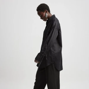 Svart långärmad skjorta från Mads Nørgaard - Säljer en stilren svart skjorta från NA-KD. Den har långa ärmar med knytning vid ärmsluten för en extra detalj. Skjortan är i ett mjukt material och har en klassisk krage. Perfekt för både vardag och fest!