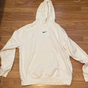 Vit hoodie från Nike - Säljer en snygg vit hoodie från Nike i storlek M. Den är i perfekt skick 10/10 med en stor Nike-logga i blått på ryggen och en mindre på framsidan. Hoodien är i standard passform och har en stor ficka framtill. Perfekt för både vardag och träning!
