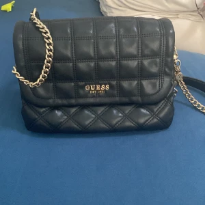 Svart quiltad axelväska från Guess - Säljer en snygg svart quiltad axelväska från Guess. Väskan har en elegant kedjerem i guld och flera små detaljer som Guess-loggan och charmiga hängen. Insidan är fodrad med Guess-mönster och har flera fack för bra organisering. Perfekt för både vardag och fest!