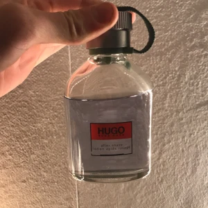 Hugo Boss after shave - Säljer en klassisk Hugo Boss after shave i en stilren glasflaska med grå kork och grön rem. Flaskan rymmer 100 ml och det är cirka 70 ml kvar. Den har en modern design med en röd etikett. Perfekt för den stilmedvetne.